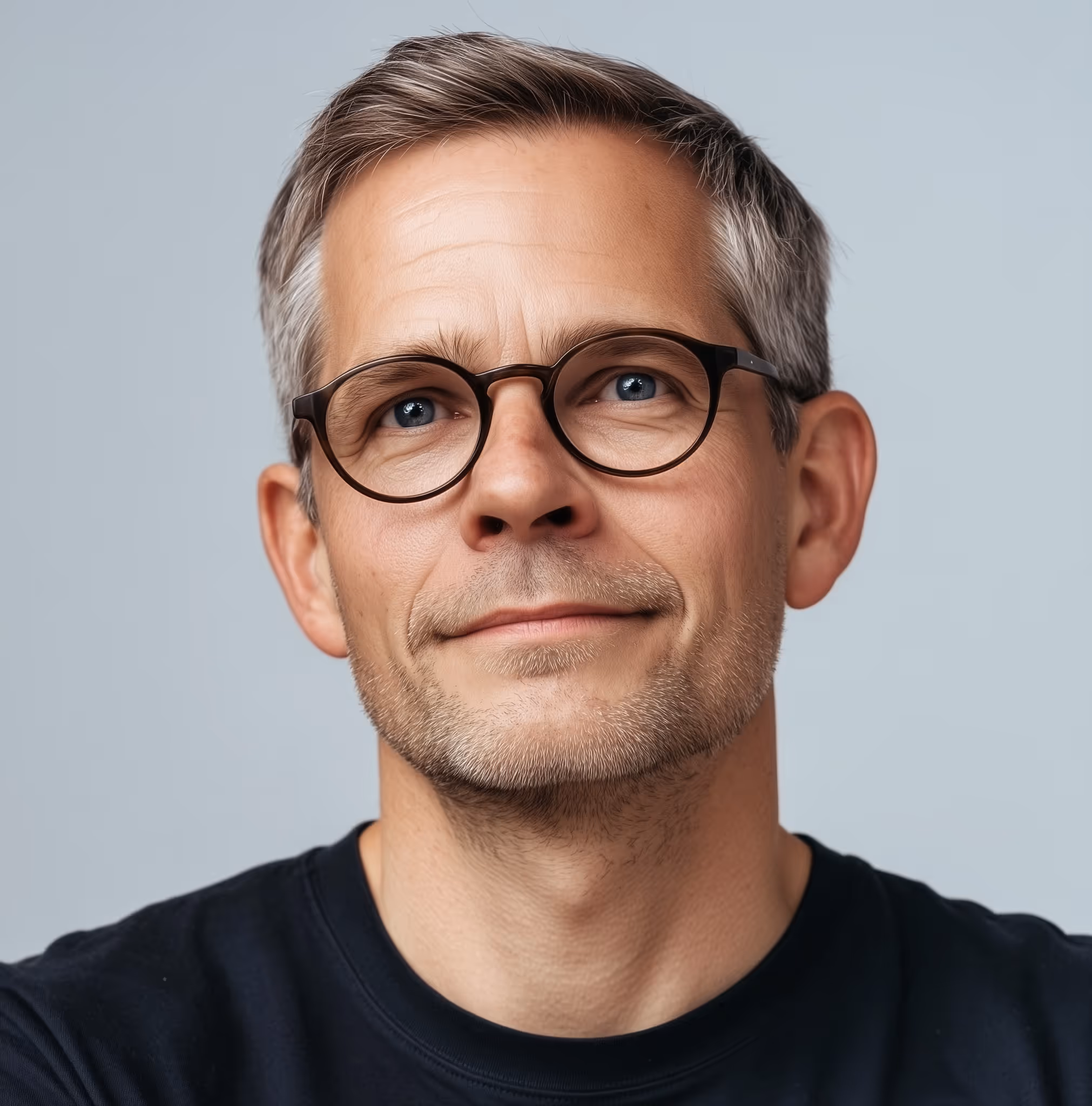 Middle-aged man with short gray hair, wearing round glasses and a black shirt, looking slightly upwards with a gentle smile.